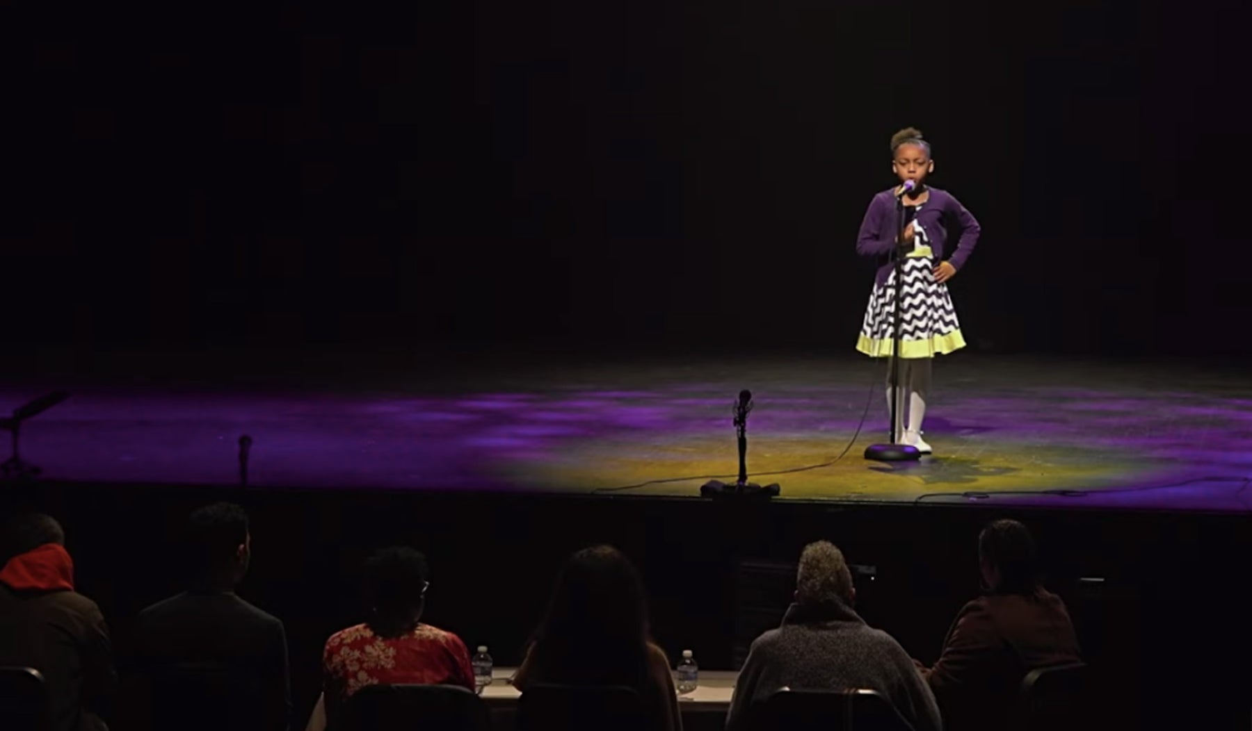 young girl in a zig zag dress on stage, speaking into mic